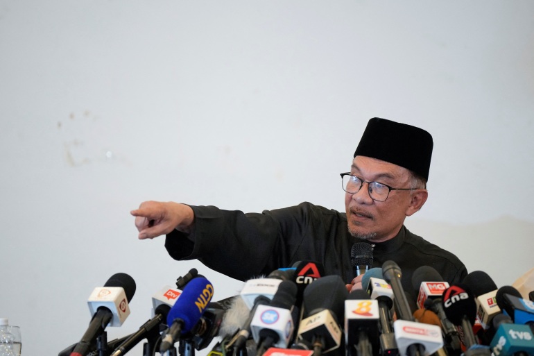 Anwar in a traditonal Malay outfit in black with songkok (black hat) points as microphones from many different networks are pushed up towards him 