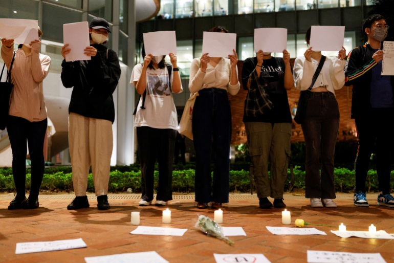 Student protesters in Hong Kong hold blank sheets of paper as part of a vigil over COVID-19 policies in China. They are wearing masks and most of their faces are obscured. There are candles lit on the floor in front of them.