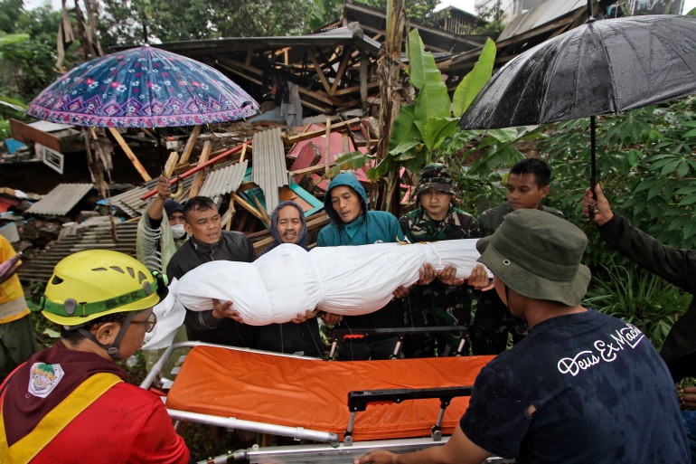 Rescuers carry out a body wrapped in a white sheet to a gurney. It is raining so one person is holding an umbrella. A collapsed house is behind