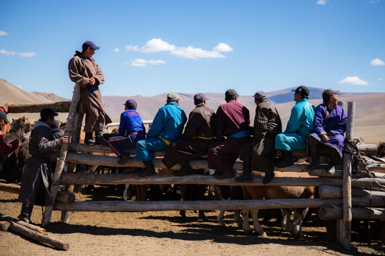 Tsaikhir Valley herders at a horse event. They are leaning over a fence with their backs to the camera. The sky is a bright blue and the landscape brown.
