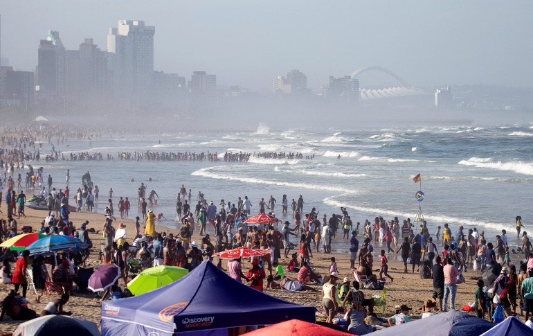 The beach is packed and there are some umbrellas and tents up. The buildings of Durban city can be seen as if in fog in the background.