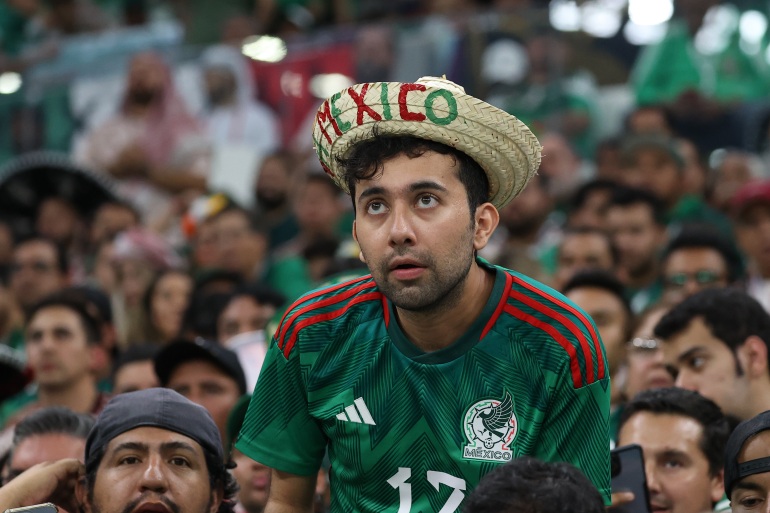 A Mexico fan wearing the team shirt and a straw hat with Mexico written in red and green on the upturned brim watches the match. He looks tense