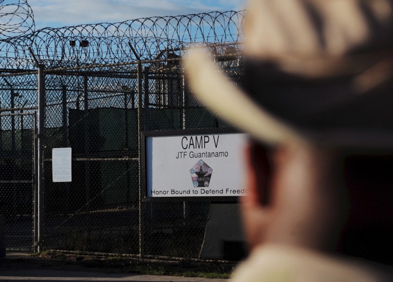 The outside of the Guantanamo Bay naval facility, with barbed wire running along the top of the fence and a sign that says: 'CAMP V, JTF Guantanamo, Honor Bound to Defend Freedom'