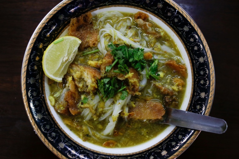 A bowl of mohinga soup, a popular breakfast for people in Myanmar. The dish contains noodles and fish. There is a slice of lime on the side and green herbs scattered on top. 