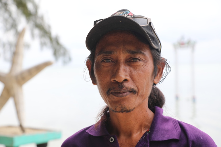 A portrait of Arif Pujianto. He has a moustache. He is wearing a purple polo shirt and a baseball cap and has perched sunglasses on the peak. He looks relaxed