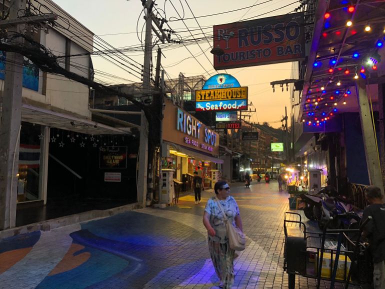 Woman walks past bar with blue, red and yellow fairy lights and a sign that says 'Russo Touristo Bar'. The street is quiet and it looks like dusk. Behind her, on the other side of the road is a large lit up sign saying 'Steakhouse'