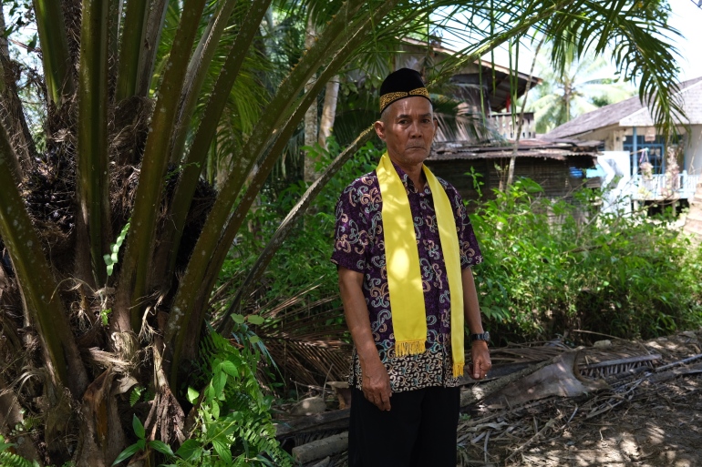 Sibukdin standing near an oil palm tree with houses behind him. He is wearing a patterned shirt and with a yellow scarf with tassles at the end draped around his neck. He is also wearing a traditional black hat with gold embroidery.